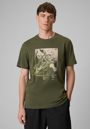 Mężczyzna w oliwkowozielonej koszulce ze wzorem abstrakcyjnej mapy topograficznej w jasnoniebieskim i beżowym kolorze, stoi na jednolitym szarym tle.