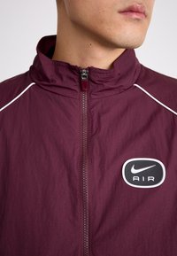 Maroon vindjacka med hög krage, vita accenter längs axlarna, framficka med dragkedja och en svart Nike AIR-logotyp på bröstet.