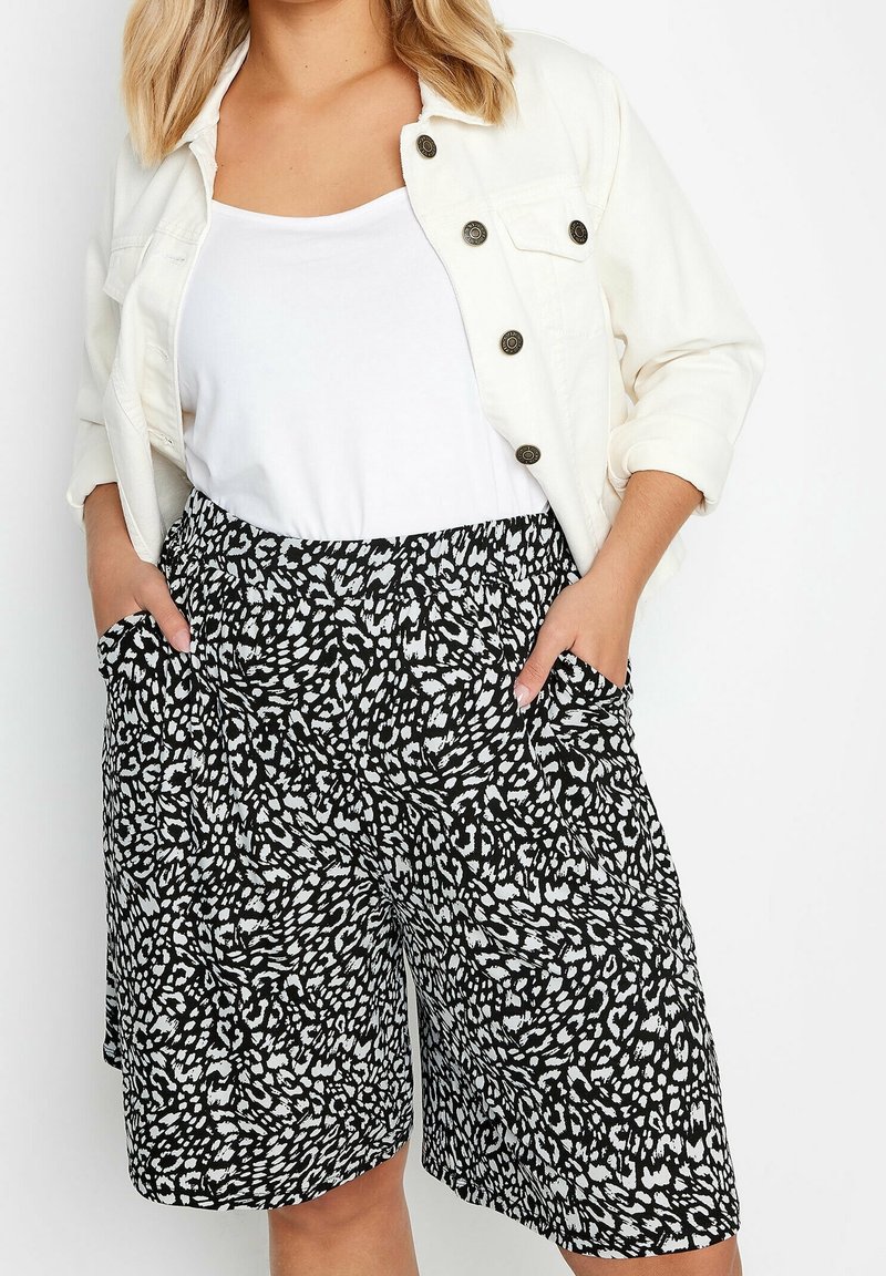 Femme portant une veste blanche, un haut blanc et un short large à motifs noir et blanc, les mains dans les poches.