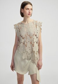 Top en crochet beige avec des motifs de feuilles et des bords festonnés, superposé à une robe légère beige lisse avec un design enveloppant.