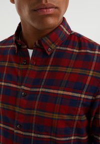 Punainen ruudullinen flanellipaita, jossa on tummansinisiä ja keltaisia yksityiskohtia, varustettuna nappikauluksella ja rintataskulla. Pehmeä rakenne ja näkyvät ompeleet.