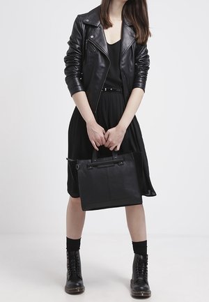 Femme portant une veste en cuir noire, une robe noire et des bottes, tenant un sac à main noir sur un fond blanc uni.