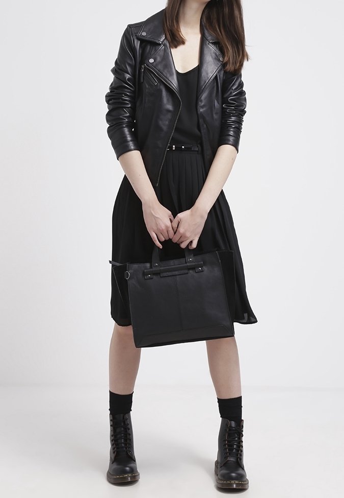 Femme portant une veste en cuir noire, une robe noire et des bottes, tenant un sac à main noir sur un fond blanc uni.
