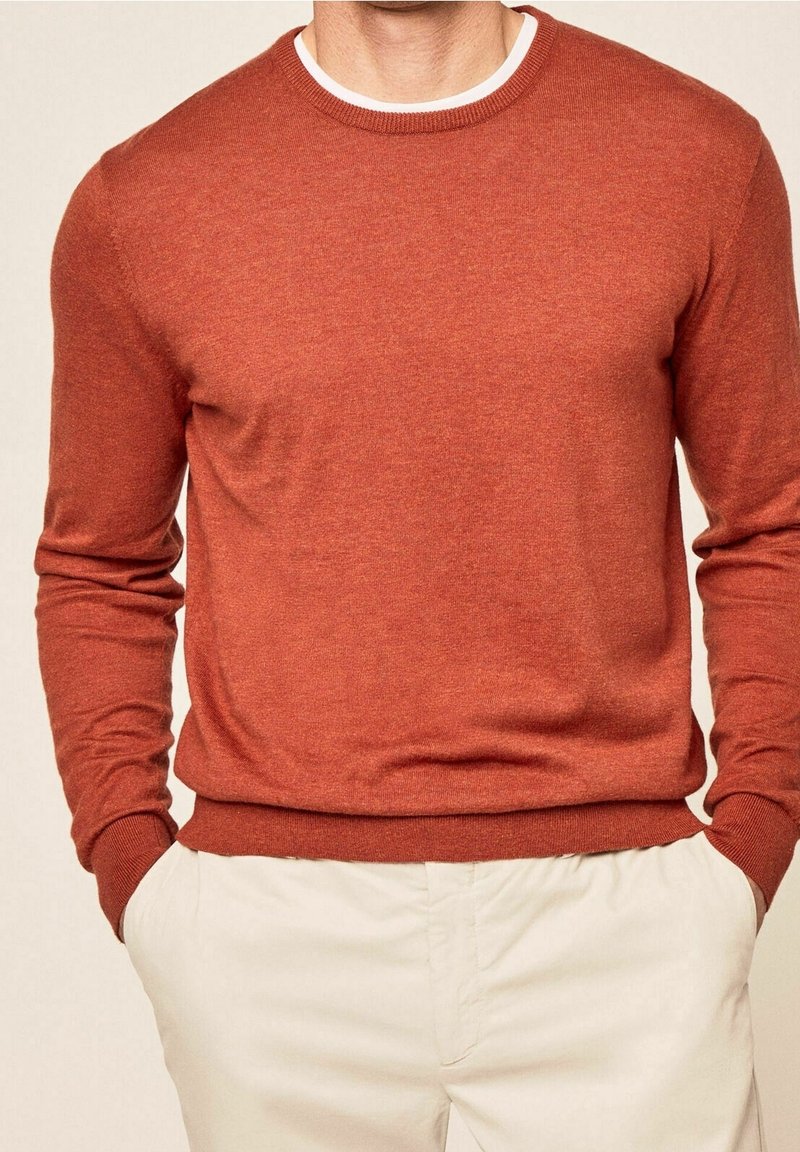 Homme portant un pull à manches longues couleur rouille et un pantalon beige clair, debout les mains dans les poches devant un fond neutre.