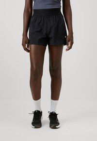 Musthave mustad lühikesed püksid laia elastse vöökohtaga, kerge kangas ja sile tekstuur, koos mustade jooksujalatsite ja valgete sokkidega.