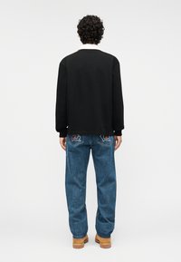 Μαύρο πουκάμισο με μακριά μανίκια, σε άνετη γραμμή, συνδυασμένο με ανοιχτό μπλε τζιν παντελόνι που έχει πίσω τσέπες με κόκκινη και μαύρη κέντηση. Καφέ παπούτσια.