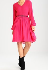 Světlý růžový plisovaný šat s dlouhými rukávy, véčkovým výstřihem a tenkým černým páskem v pase, doplněný černými kotníkovými botami.