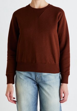 Personne portant un sweat-shirt marron foncé uni à manches longues et un jean bleu clair délavé sur un fond blanc uni.
