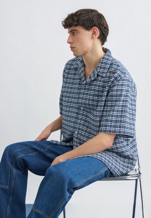 Jeune homme aux cheveux bouclés courts portant une chemise à manches courtes à carreaux bleus et blancs et un jean bleu, assis de côté sur une chaise en métal.