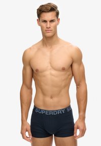Boxers noirs en bleu foncé fabriqués en tissu doux, avec une large ceinture élastique arborant le logo "SUPERDRY" en gris. Texture lisse et design ajusté.