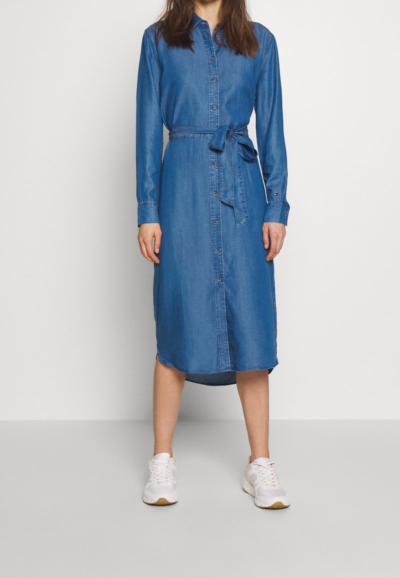 Robe chemise en denim, devant boutonné, manches longues, taille cintrée avec un lien, de couleur bleu uni, longueur genoux, associée à des sneakers blanches.