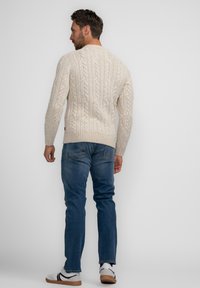 Cremfarbener Kabelstrickpullover mit gerippten Bündchen, kombiniert mit blauen Jeans und weißen Sneakers. Der Pullover hat ein strukturiertes Muster.