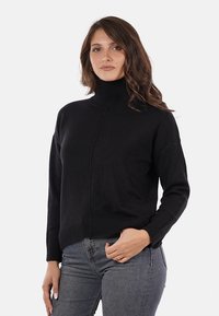 Donna con capelli castani ondulati, che indossa un maglione nero a collo alto e jeans grigi, in piedi contro uno sfondo chiaro e semplice.