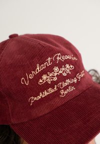 Burgundy bordás sapka bézs hímzett virágmintával és a "Verdant Reverie Prohibited Clothing Berlin" felirattal az elején.