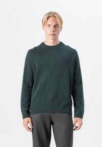 Pulover tricotat de culoare verde închis, cu guler rotund, mânici și tiv cu nervuri, asortat cu pantaloni gri. Textile moale cu un design minimalist.