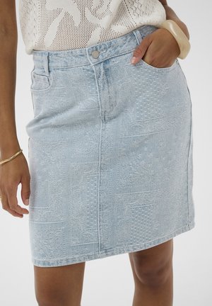 Svetlomodrá texturovaná rifľová sukňa nosená s krémovým pleteným topom, ruka v vrecku, doplnená béžovým náramkom a tenkým zlatým náramkom.