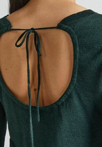 Zielony dziergany sweter z okrągłym tyłem, dużym owalnym wycięciem oraz zawiązywaniem, wykonany z miękkiego, teksturowanego materiału.
