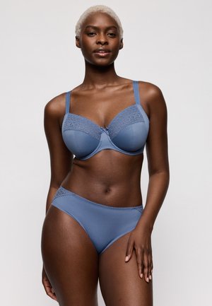 Vrouw met kort blond haar die een bijpassende blauwe kanten beha en hoog uitgesneden ondergoed draagt, staand tegen een eenvoudige lichte achtergrond.