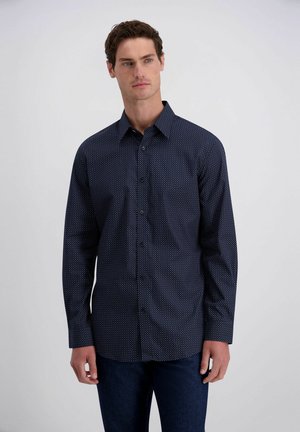Man draagt een donkerblauw overhemd met lange mouwen en kleine witte stippen, staand tegen een eenvoudige lichte achtergrond.