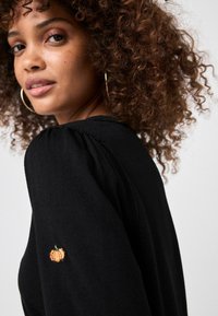 Maglione nero a maglia con maniche a palloncino, caratterizzato da un piccolo ricamo di una zucca arancione sulla manica. Morbido al tatto e dal design aderente.