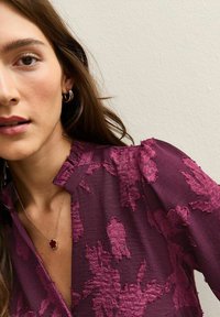 Burgundowa bluzka z kwiecistą fakturą i falbaniastym dekoltem. Naszyjnik w odcieniu złota z czerwonym wisiorkiem podkreśla całość.