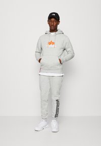 Felpa grigia con logo arancione, pantaloni della tuta grigi abbinati con stampa nera e sneakers bianche. Il modello indossa un cappellino nero. Design semplice, stile casual.