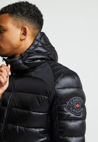 Čierna prešívaná bunda s vysokým golierom a nastaviteľnou kapucňou, ktorá má nášivku loga na ľavom rukáve a prešívanú štruktúru.