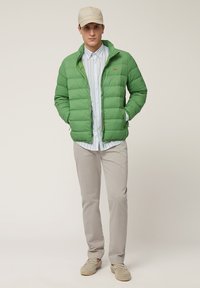 Giacca puffer verde con sezioni trapuntate, chiusura con zip e logo rosso. Indossata sopra una maglietta a righe, abbinata a pantaloni beige e scarpe slip-on.