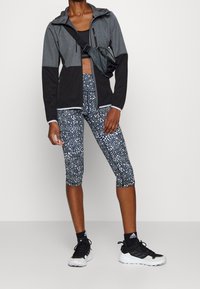 Veste zippée grise et noire avec capuche, superposée à un soutien-gorge de sport noir ; leggings capri à motifs bleu et blanc ; chaussures de sport noires.