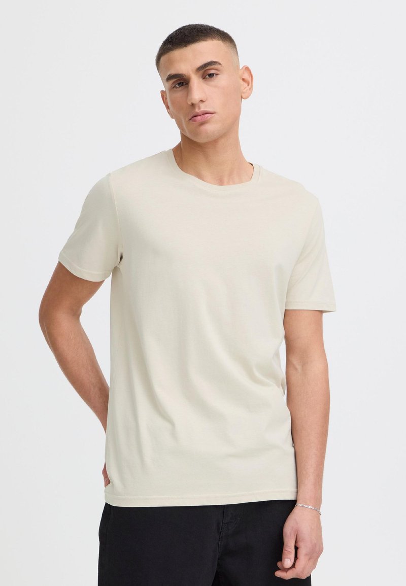 Junger Mann mit kurzen Haaren, der ein schlichtes hellbeiges T-Shirt und schwarze Hose trägt, steht vor einem weißen Hintergrund.