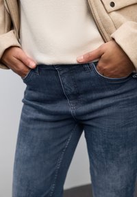 Jeans ajustados azul oscuro con un acabado texturizado, presentan bolsillos frontales y un diseño de cinturilla simple, usados debajo de una blusa clara.