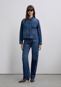 Denimjakke med farverige udsmykninger, kombineret med vidde bukser i denim. Mørkeblåt stof, knapdetaljer og let slid ved kanten.