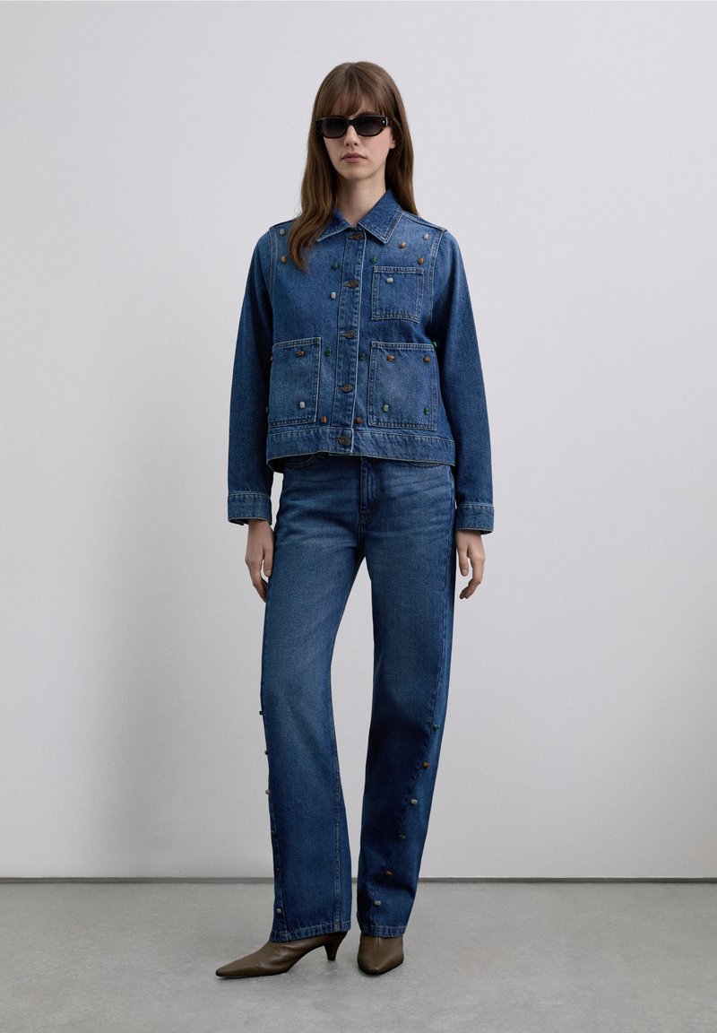 Denimjakke med farverige udsmykninger, kombineret med vidde bukser i denim. Mørkeblåt stof, knapdetaljer og let slid ved kanten.