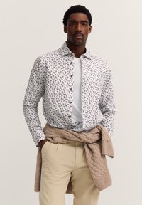 Ein Mann trägt ein weißes, gemustertes Hemd über einem weißen T-Shirt, beigefarbene Hosen und einen beigefarbenen, grob gestrickten Pullover um die Taille gebunden.