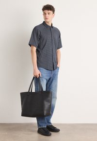 Μαύρη δερμάτινη τσάντα ώμου με διπλές λαβές, ορθογώνιο σχήμα, λεία επιφάνεια, τοποθετημένη σε ουδέτερο φόντο με ανδρικό μοντέλο.