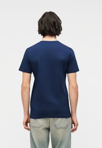 Άνδρας με μεσαίου μήκους σκούρα μαλλιά, φορώντας ένα απλό κοντομάνικο μπλε σκούρο μπλουζάκι και ανοιχτόχρωμα τζιν παντελόνια, γυρισμένος προς τα πίσω πάνω σε λευκό φόντο.