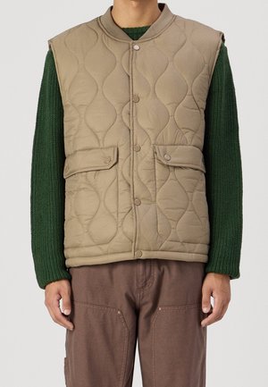 Man wearing a beige quilted vest with snap buttons over a dark green knit sweater and brown pants.