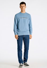 Ljusblå sweatshirt med texten "KÖPENHAMN", rund halsringning och ribbade mudd, i kombination med mörkblå jeans och svarta sneakers.