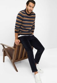 Multicolored gestreepte trui in bruin, blauw en crème, gecombineerd met donkere corduroy broek en witte sneakers. Model zit op een houten stoel.