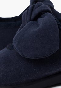 Scarpa in tessuto blu scuro con un grande e morbido fiocco sulla parte superiore. Presenta una superficie testurizzata e una suola bassa e robusta.