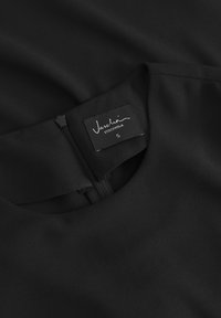 Crna haljina s okruglim izrezom, patentnim zatvaračem na leđima i tekstilnom etiketom koja identificira marku kao Vascha. Veličina S.