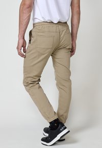 Pantaloni jogger bej cu manșete și talie elastice, având două buzunare la spate și un buzunar lateral cu fermoar, asortați cu teniși negri.