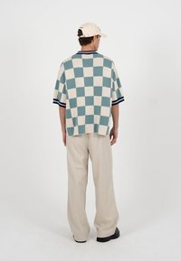 Mand iført cremefarvet kasket, blå og cremefarvet ternet kortærmet sweater, vide beige bukser og sorte sko, stående med ryggen mod en hvid baggrund.