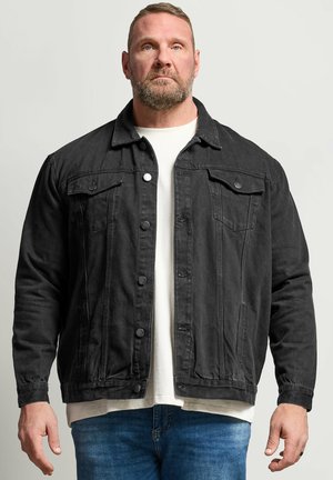 Man met baard die een zwarte spijkerjas draagt over een wit overhemd en blauwe spijkerbroek, staand en recht naar voren kijkend tegen een effen achtergrond.