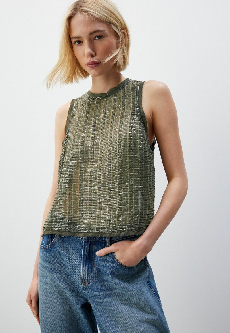 Mouwloos olijfgroen topje met een geweven stof met verticale strepen en pailletten. Ruwe rand halslijn; gecombineerd met blauwe jeans.
