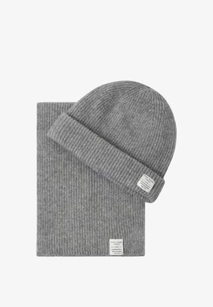 Grijze geribbelde gebreide beanie en bijpassende sjaalset, elk met een klein wit merkje genaaid aan de rand.