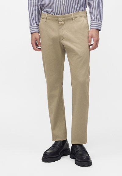 Homme portant un pantalon beige coupe droite, des mocassins noirs en cuir et une chemise à rayures bleues et blanches boutonnée, debout devant un fond blanc.