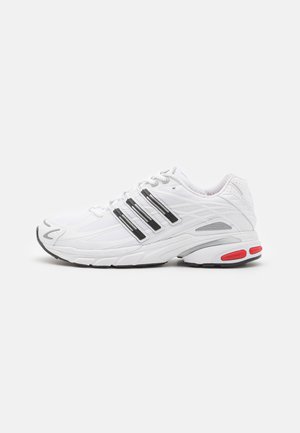 Biały sportowy sneaker z czarnym wzorem trzech pasków, teksturowaną podeszwą, szarymi akcentami i czerwonym detalem na pięcie na jednolitym tle.