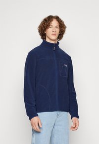 Giacca in pile blu marina con colletto alto, chiusura frontale a zip completa e patch con logo. Presenta tasche laterali e un tessuto morbido e texturizzato.
