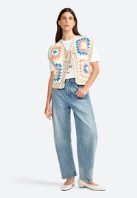 Crochet vest in cream with blue and pink granny squares, tied at the center. Paired with a plain white t-shirt and light blue wide-leg jeans.